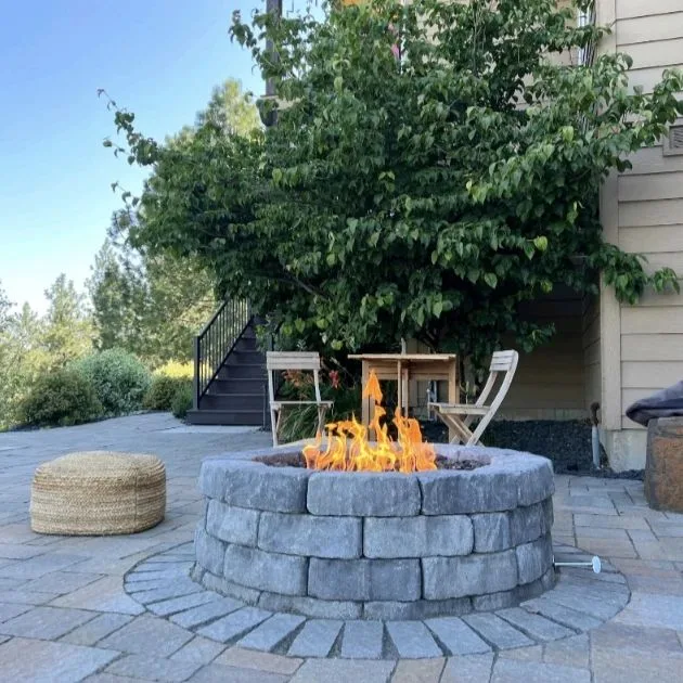 Round paver gas fire pit burning beside bistro chairs on a backyard patio