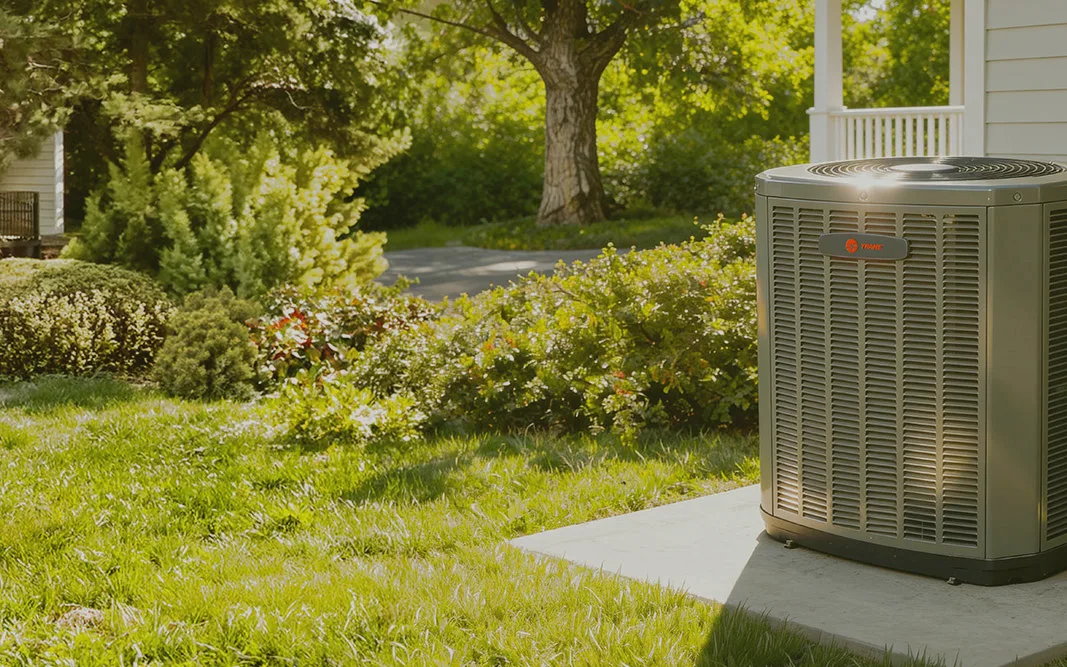 A Trane outdoor unit on its pad beside a white-sided home, with a lawn and shrubs